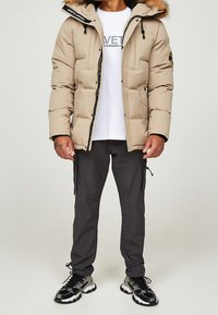 Chaqueta acolchada beige con capucha de piel sintética, cierre con cremallera y dos bolsillos frontales. Llevada sobre una camiseta blanca con logo gris, combinada con pantalones oscuros y zapatillas.