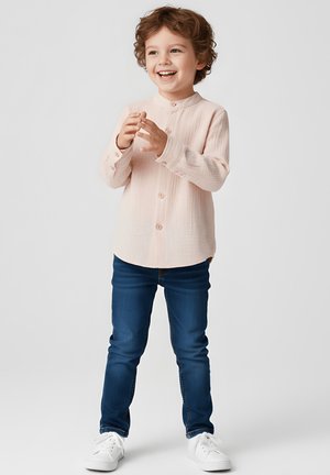 Jasnoróżowa koszula na guziki z kołnierzykiem typu mandarynka, zestawiona z ciemnoniebieskimi dżinsami i białymi trampkami. Materiał ma teksturowany wygląd.