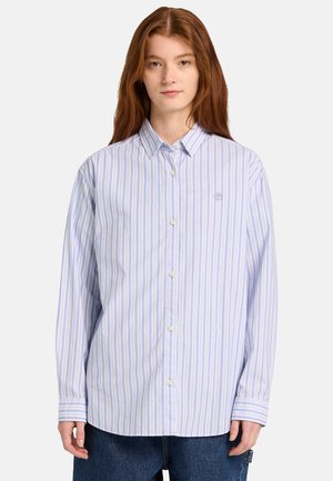 Ung person med langt rødt hår iført en lyseblå og hvid stribet skjorte med knapper og mørkeblå jeans, set forfra.