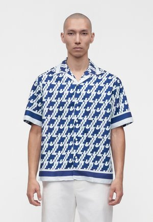 Homme portant une chemise à manches courtes boutonnée avec un motif géométrique bleu et blanc et un pantalon blanc, debout devant un fond uni.