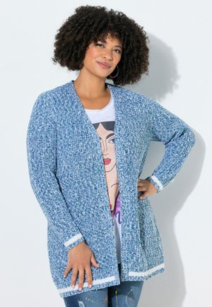 Kvinde med krøllet hår iført blå strikket cardigan over en graphic t-shirt, stående med den ene hånd på hoften foran en hvid baggrund.