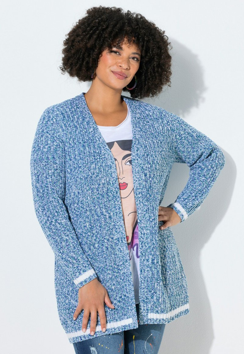 Femme aux cheveux bouclés portant un cardigan en maille bleu sur un t-shirt graphique, debout avec une main sur la hanche, sur fond blanc.