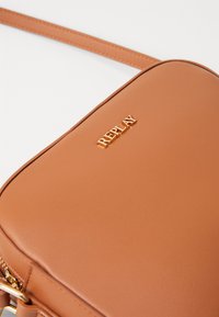 Lærcrossbody-veske i tan med en jevn tekstur, rektangulær form, og en gull "REPLAY" logo preget på fronten. Justerbar stropp inkludert.
