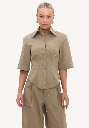 Femme aux cheveux blonds porte une chemise beige à manches courtes boutonnée et un pantalon assorti orné de petits rivets métalliques, debout devant un fond uni.
