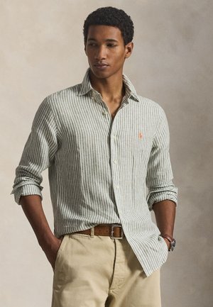 Mann trägt ein grün-weiß gestreiftes Langarmshirt, beige Hose, braunen Gürtel und Uhr, steht mit Händen in den Taschen vor neutralem Hintergrund.