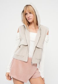 Μπεζ καπιτονέ γιλέκο με κουκούλα και δύο μπροστινές τσέπες, φορεμένο πάνω από ένα λευκό τοπ. Το μοντέλο το συνδυάζει με μία ροζ φούστα με υφή.