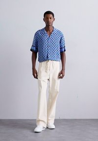 Chemise à manches courtes bleue à motifs avec une coupe décontractée, associée à un pantalon crème léger et des baskets blanches, se tenant devant un fond neutre.