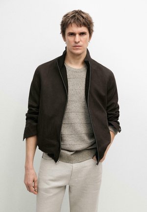 Mann trägt eine dunkelbraune Zip-up-Jacke über einem beigen Strickpullover und hellen Hosen, steht mit einer Hand in der Tasche vor einer weißen Wand.