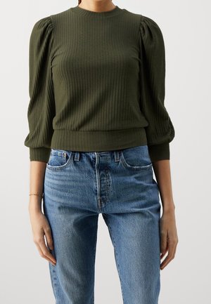 Vrouw met een olijfgroen geribd langärmelig shirt met pofmouwen en een high-waisted blauwe spijkerbroek tegen een effen achtergrond.