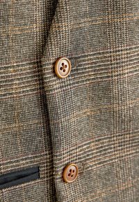 Braun karierter Blazer aus strukturiertem Stoff, mit zwei braunen Knöpfen und einem schwarzen Taschenbesatz.