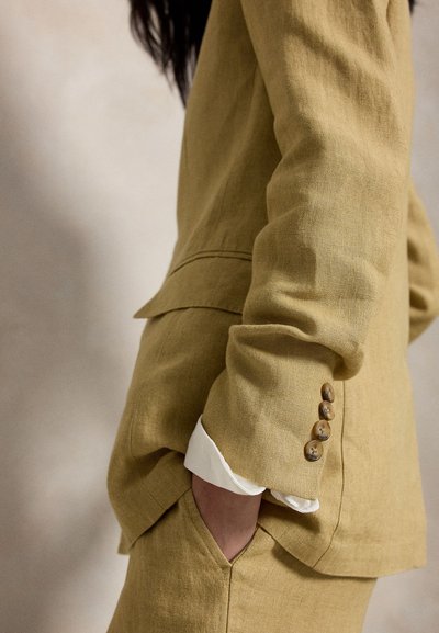 Inimene kannab sinepikollast linast ülikonda, mille varrukal on neli nööpi, valge särgivarukas paistab, käsi on pükstaskus neutraalse tausta ees.