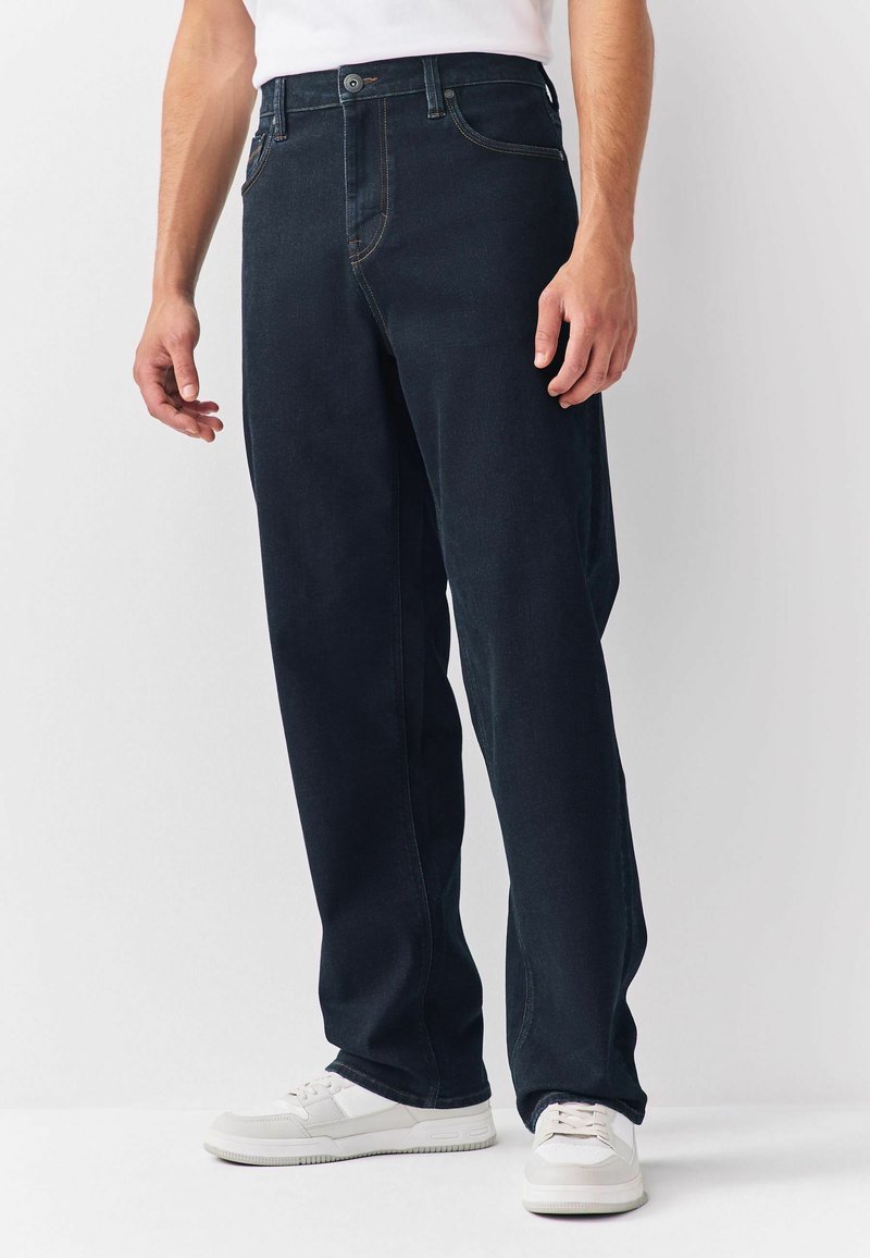 Mand iført mørkeblå lige ben jeans og hvide sneakers, stående mod en ensfarvet lys baggrund.