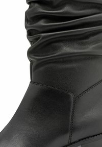 Prim-plan al unei cizme din piele neagră cu un branț moale, adunat și cusături detaliate vizibile pe suprafața texturată.