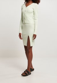 Světle zelený žebrovaný kardigan s výstřihem do V, zapínáním na knoflíky a ladící sukně se bočním rozparkem. Černé sandály dotvářejí celý outfit.