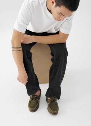 Νεαρός με κοντά μαλλιά που κάθεται σε ένα κουτί, φορώντας λευκό T-shirt, μαύρα τζιν, παπούτσια ελιάς και τατουάζ στον αντιβράχιο.