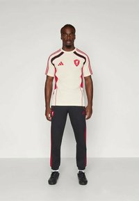 Weißes und schwarzes Sportshirt mit roten Akzenten und Logo, kombiniert mit schwarzen Hosen mit roten Streifen, getragen mit schwarzen Schuhen.