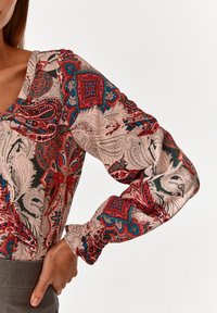 Blouse à manches longues en tissu léger avec des motifs paisley et floraux complexes dans des teintes de rouge, bleu et crème. Elle présente un décolleté en V et des poignets froncés.
