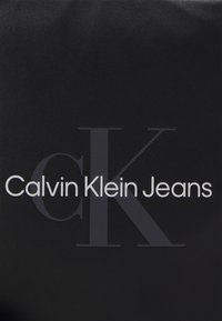 Borsa in pelle nera con il testo "Calvin Klein Jeans" in bianco e un grande logo "CK" grigio, texture liscia, design minimalista.