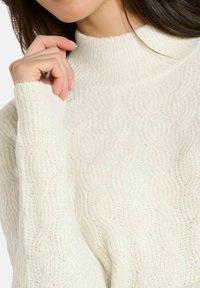 Cremfarbener, gestrickter Pullover mit einem strukturierten Wellenmuster, hochgeschlossenem Kragen und gerippten Manschetten. Weiches, leicht schimmerndes Material.