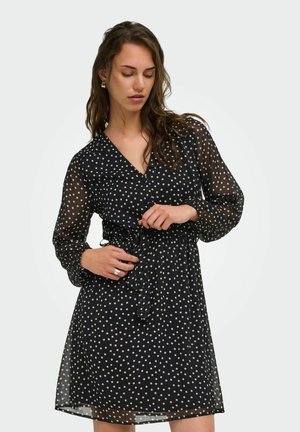 Femme ajustant la ceinture d'une robe noire à pois blancs avec des manches longues transparentes, debout devant un fond clair uni.