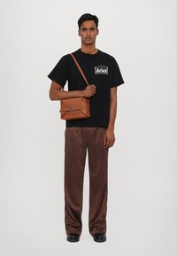 Black t-shirt with white logo, brown satin trousers, brown crossbody bag with clasp, and black shoes. Model stands against a white background.
