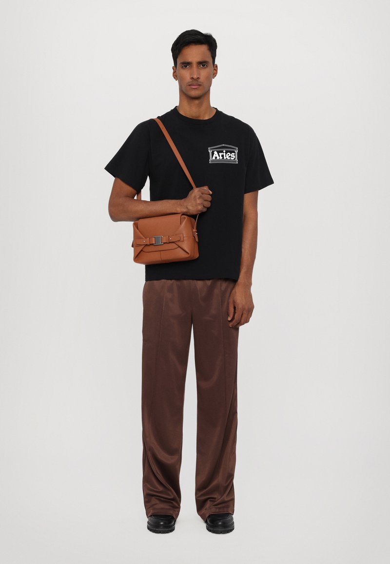 Black t-shirt with white logo, brown satin trousers, brown crossbody bag with clasp, and black shoes. Model stands against a white background.