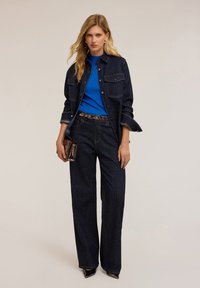 Giacca in denim scuro e jeans a gamba larga con motivo a pois, abbinati a un top blu e una clutch lucida. Tessuto testurizzato, dettagli di cucitura discreti.