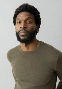 Olijfgroene, getailleerde lange mouwen shirt met een ronde halslijn. Zachte textuur, geribbelde stof, minimalistisch ontwerp. Model heeft krullend haar en een baard.