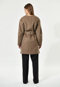 Cappotto di lana marrone con maniche ampie e vita con cintura. Presenta un taglio dritto e arriva a metà coscia, abbinato a pantaloni neri.
