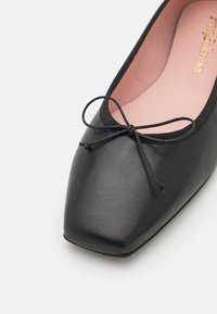 Ballerine plate en cuir noir avec bout carré et délicat nœud sur le dessus, dotée d'une semelle intérieure rembourrée rose clair.