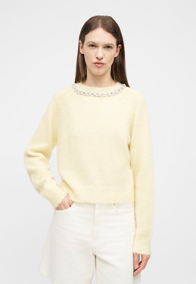 Γυναίκα με ίσια καστανά μαλλιά, που φοράει ένα κρεμ πουλόβερ με λεπτομέρεια από πέρλες στη λαιμόκοψη και λευκό παντελόνι με ψηλή μέση.