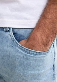 Světle modré džíny z denimu s přední kapsou, které mají měkkou texturu a viditelné švy. Ruka odpočívá v kapse.