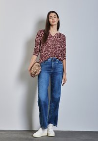 Rote und beige gemusterte Bluse mit 3/4-Ärmeln, kombiniert mit blauen weitem Beinen Jeans und weißen Turnschuhen, hält eine beige geflochtene Clutch.