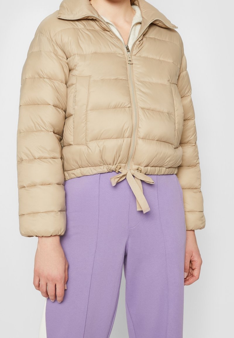 Beige puffjacka med en kort design, horisontellt kviltad textur, dragkedjestängning och dragsko i fållen. Har en klassisk krage och sidofickor.