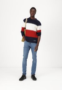 Pull en tricot avec des rayures bleu marine, crème et rouge, à col rond et avec des bords-côtes. Associé à un jean bleu clair et des baskets noires.