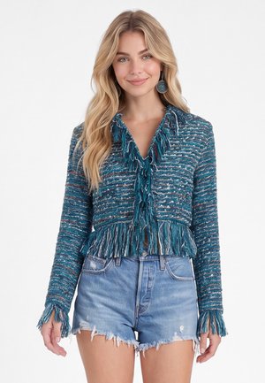 Giovane donna con lunghi capelli biondi indossa un maglione lavorato a maglia blu con frange e pantaloncini di jeans strappati, sorridendo su uno sfondo bianco.