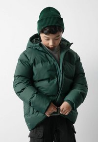 Zielona kurtka puchowa z kapturem, z pikowanym wzorem i ściągaczami na rękawach. Nosi ją osoba na szarym podkoszulku, w zestawie z pasującą zieloną czapką robioną na drutach.