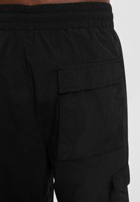 Schwarze Cargo-Hose mit elastischem Bund, glatter Textur und großer Seitentasche mit Klappdetail. Schlichtes und funktionales Design.