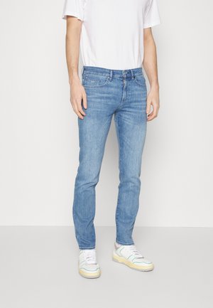 Homme portant un jean slim bleu clair, un t-shirt blanc et des baskets blanches avec des touches vertes et jaunes, debout devant un fond uni.