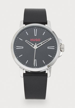 Fekete bőrszíjas óra ezüst tokkal, kerek, sötétszürke számlap, fehér órák jelölőkkel és vörös "HUGO" logóval. Texturált szíj jellemzi.