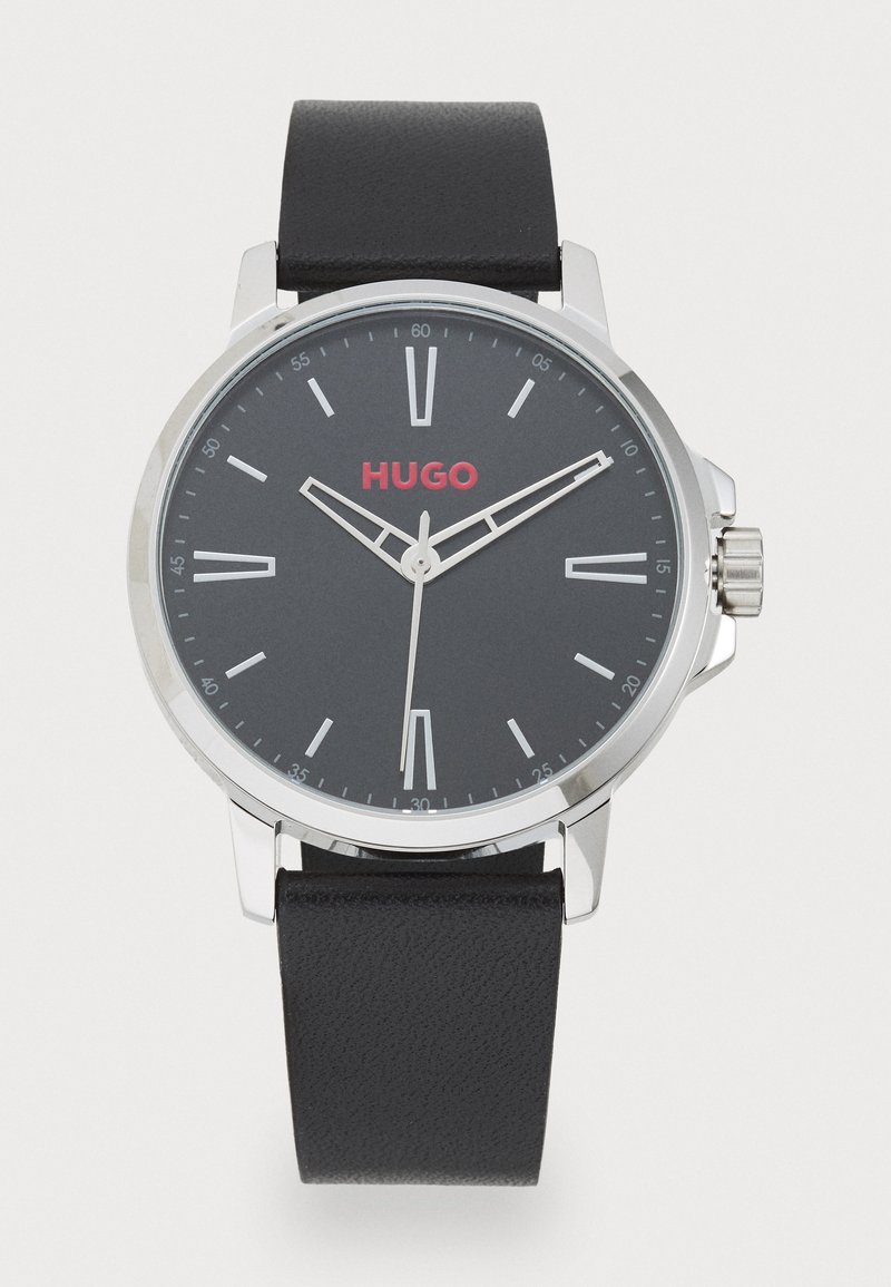 Juodasis odinis laikrodžio dirželis su sidabriniu korpusu, apvaliu tamsiai pilku ciferblatu, baltais valandų žymekliais ir raudonu "HUGO" logotipu. Oda dirželis turi tekstūruotą paviršių.
