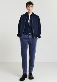 Marineblaue Bomberjacke, dunkles Hemd und marineblaue Cordhosen. Schwarze Stiefeletten runden das Outfit ab und betonen einen schlanken und modernen Look.