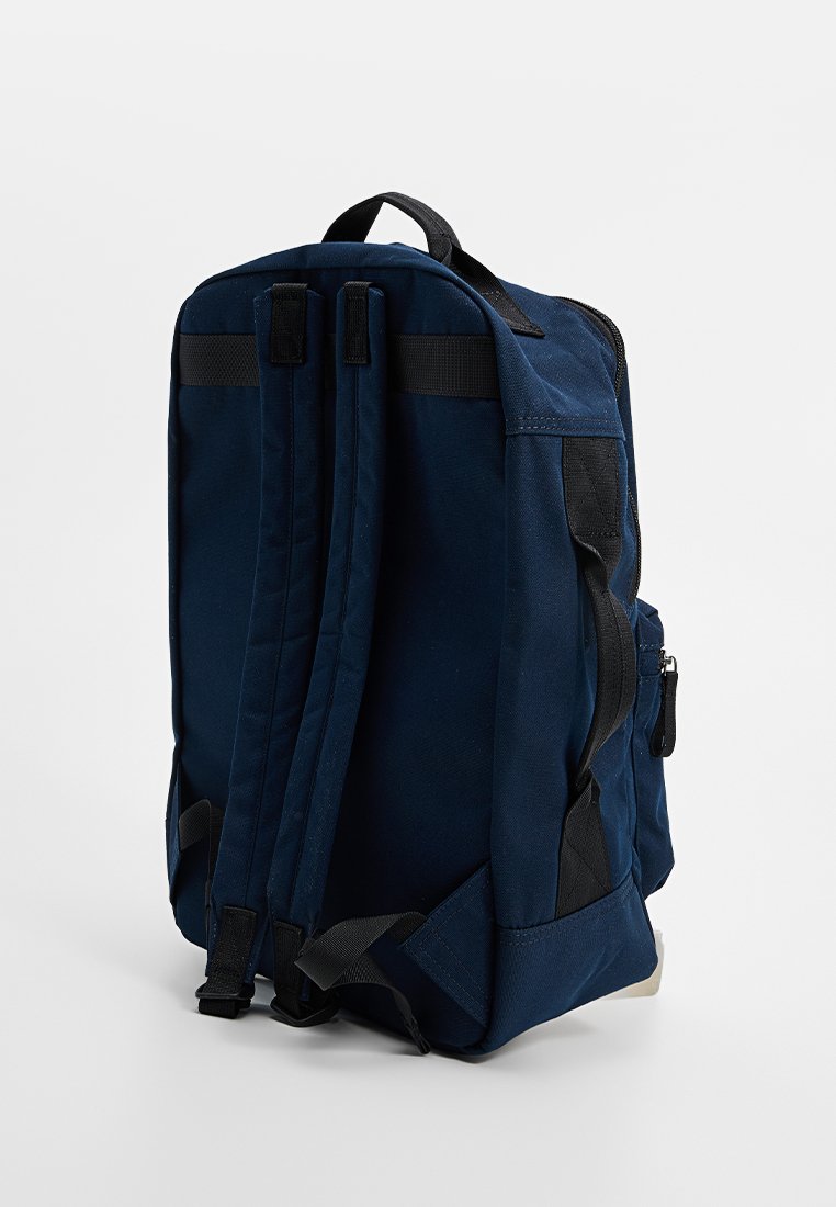 Mochila azul marino hecha de tela resistente, con correas ajustables, un bolsillo lateral y acentos negros para contraste. Forma rectangular.