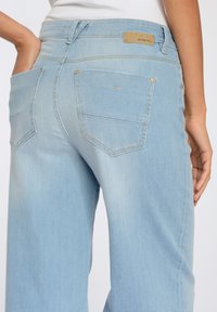 Světlé modré džíny s kapsami na zadní straně. Mají středně vysoký pas, nenápadné švy a texturovanou etiketu na pase.