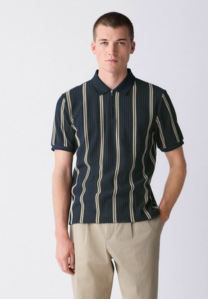 Mężczyzna ubrany w granatową koszulkę polo z pionowymi kremowymi paskami i beżowe spodnie, stojący z jedną ręką w kieszeni na białym tle.