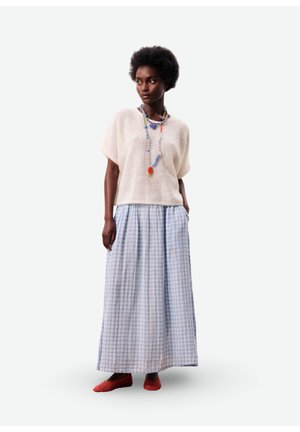 Femme portant un pull à manches courtes blanc, une longue jupe à carreaux bleue et blanche, un collier coloré de perles, et des ballerines rouges, debout les mains dans les poches.