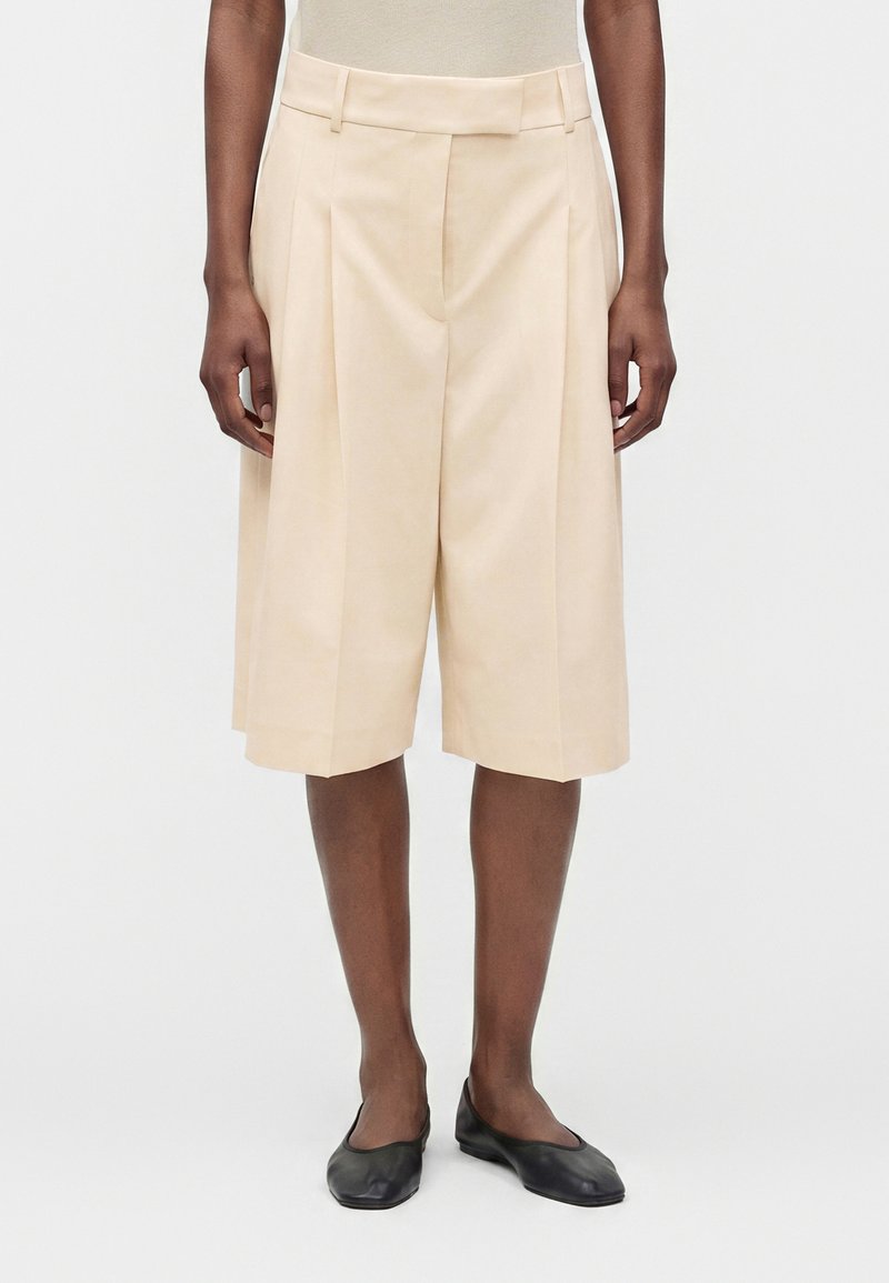 Personne portant un short beige sur mesure mi-long avec des plis, un haut blanc et des chaussures plates noires, debout sur un fond blanc.