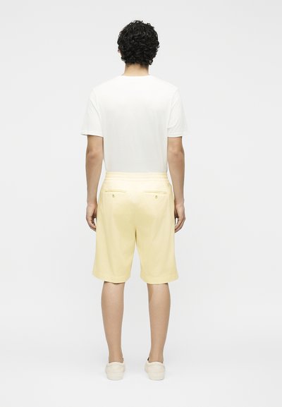 Άνδρας με σγουρά μαλλιά που φοράει λευκό μπλουζάκι, ανοιχτόκκινα κίτρινα σορτς μέχρι το γόνατο με πίσω τσέπες και λευκά παπούτσια slip-on, στέκεται μπροστά σε λευκό φόντο.