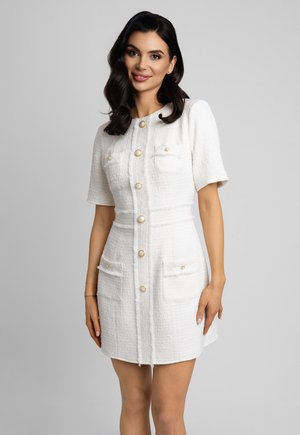 Kobieta o ciemnych, falowanych włosach, ubrana w białą, teksturowaną sukienkę z krótkimi rękawami, ozdobioną złotymi guzikami i przednimi kieszeniami, stojąca na tle szarego tła.
