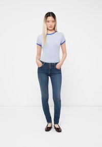 Γυναίκα που στέκεται με τα χέρια στις τσέπες, φορώντας μπλε και λευκή ριγέ μπλούζα, σκούρα στενά τζιν και μπορντό φλατ παπούτσια, μπροστά σε λευκό φόντο.